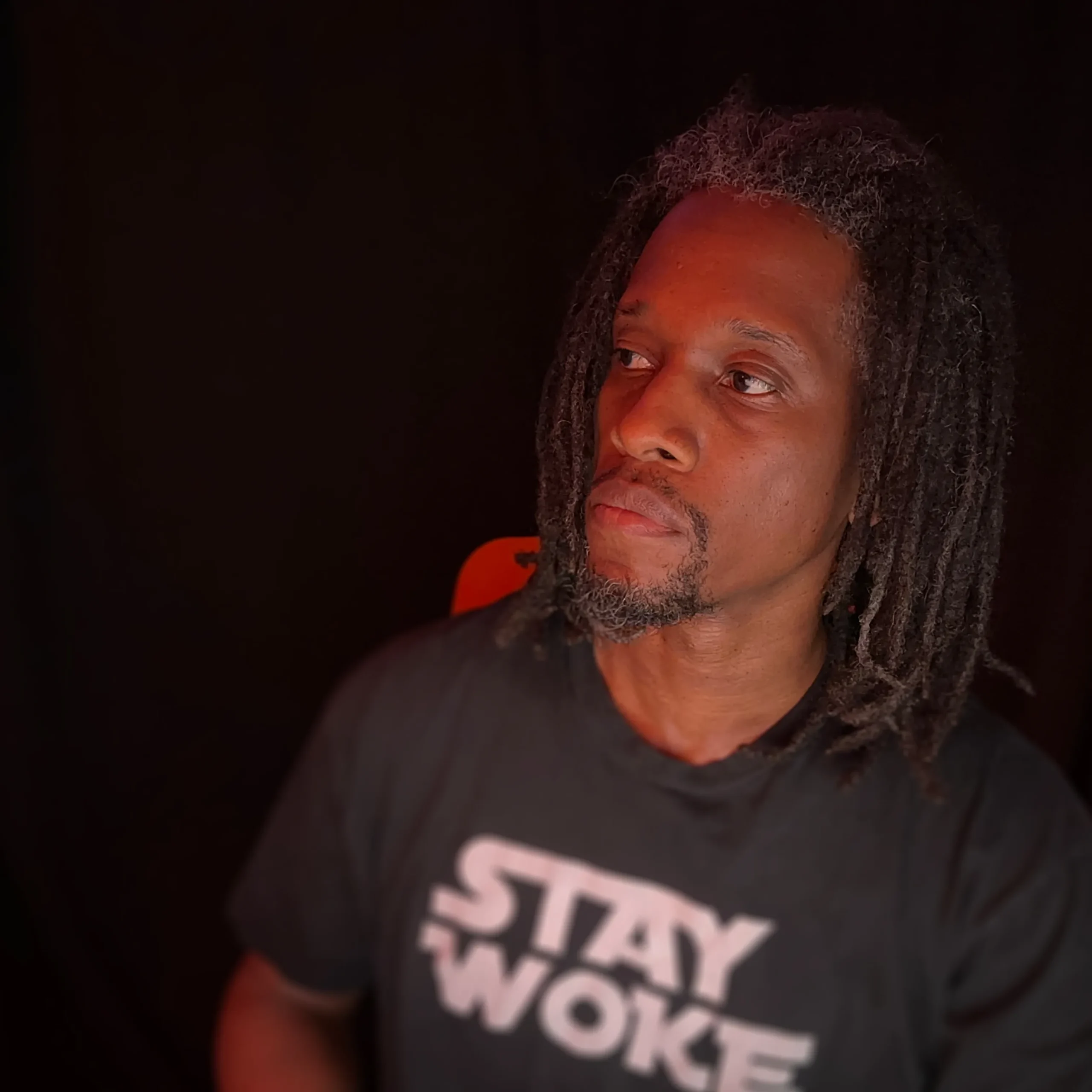 Q. looking off to his right, with a black t-shirt that says "Stay Woke," in front of a black backdrop.