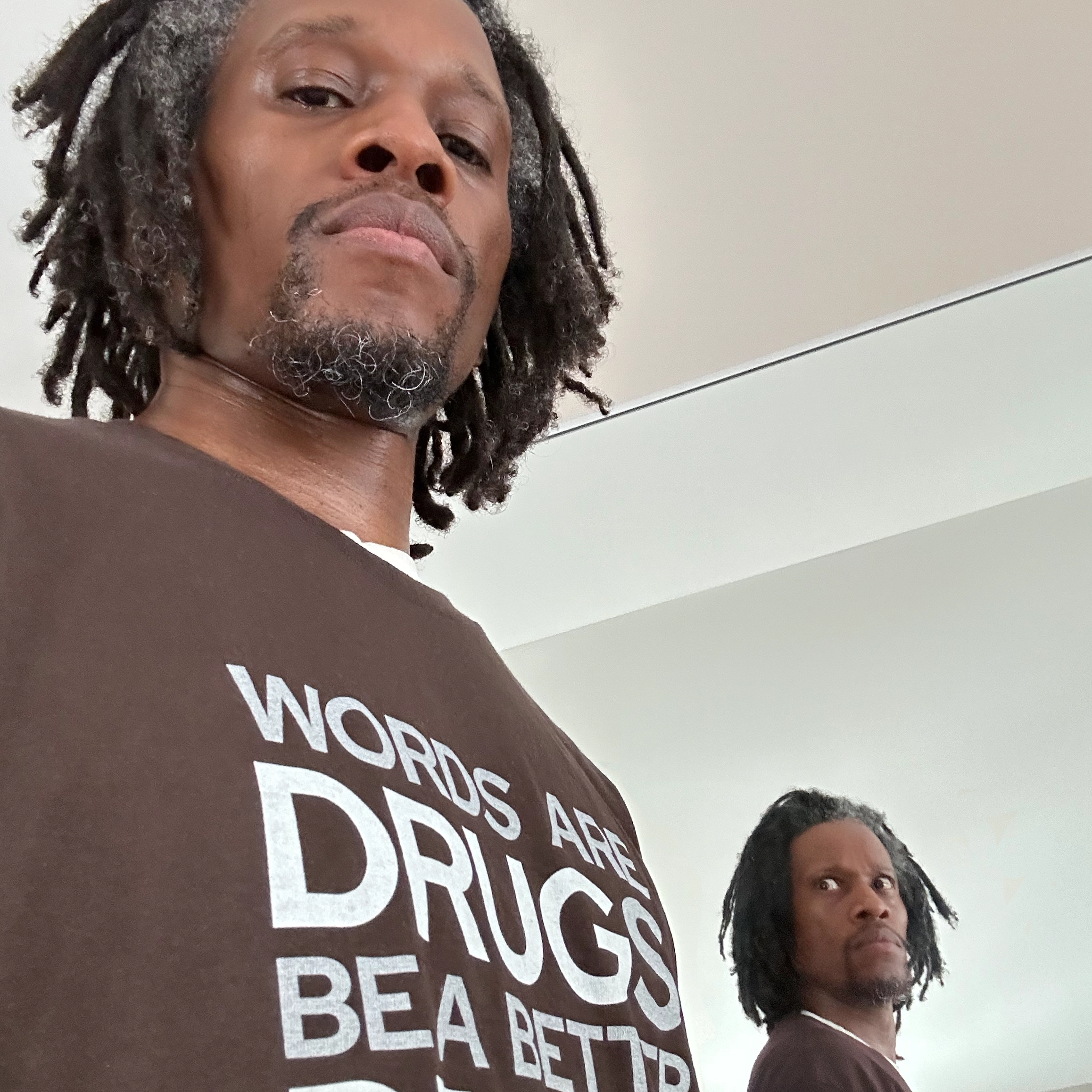 Photo of Q standing in front of a mirror, in a brown shirt that says "Words are drugs, be a better dealer," with the image of himself in the mirror looking back at him menacingly.